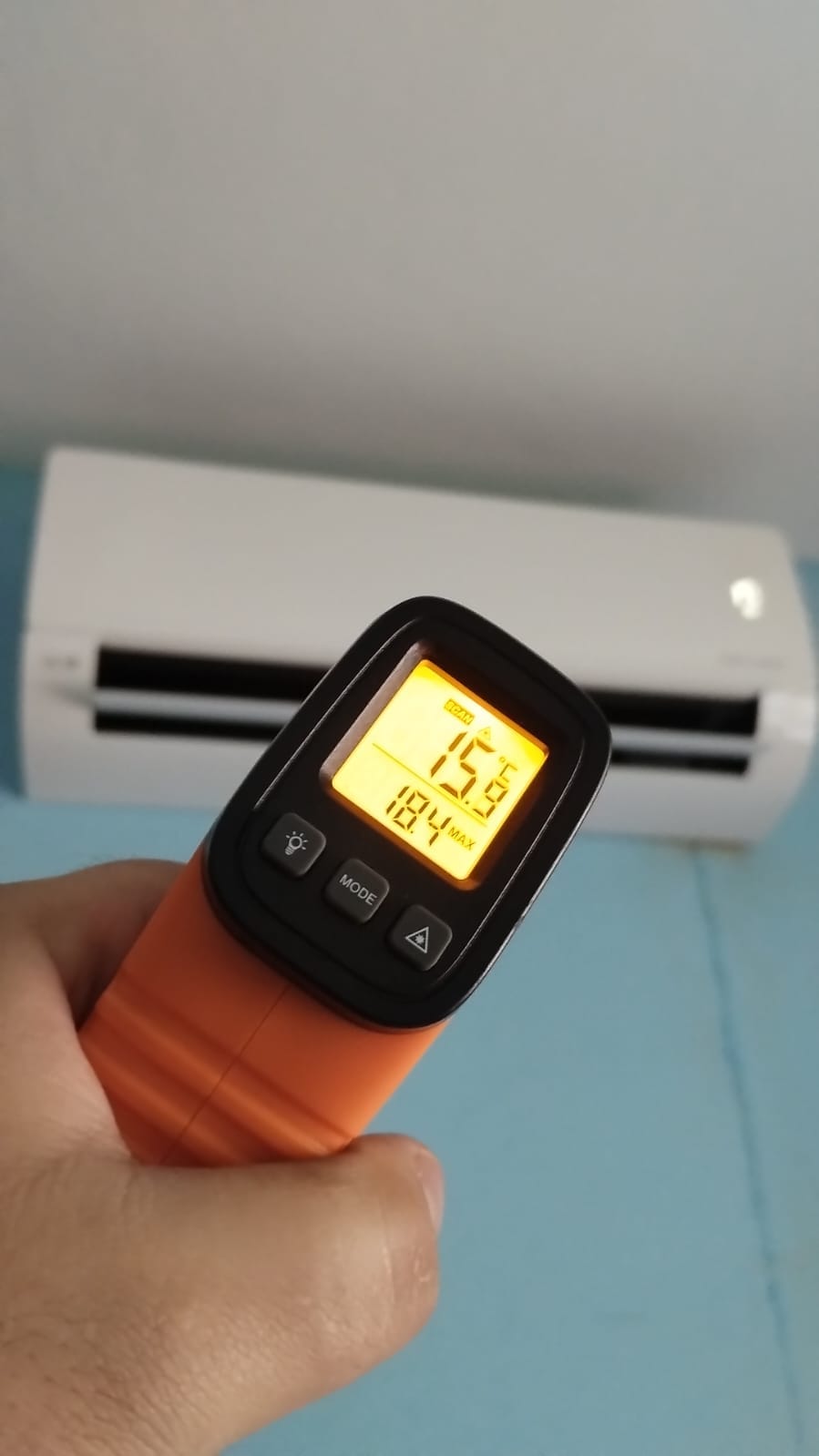 Mão segurando termômetro infravermelho laranja marcando 15.8°C na frente do ar-condicionado split branco